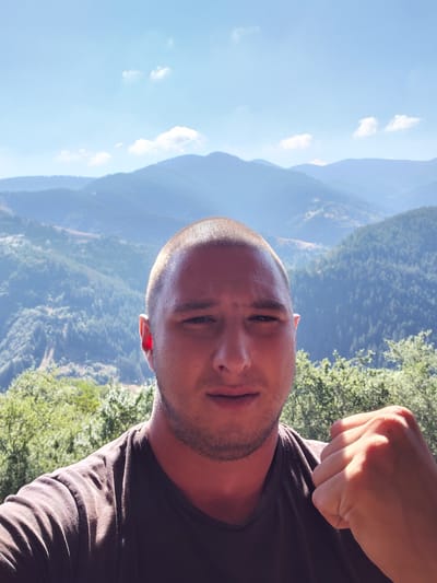 A close-up selfie shows a man with a shaved head and a beard standing outdoors. He is wearing a dark t-shirt and his right fist is clenched and raised towards the camera. The background is a panoramic view of lush green mountains and a clear blue sky with scattered clouds, suggesting the image was taken in a natural, mountainous area. The specific location in Dryanovets, Bulgaria, is not discernible from the visual elements in the image itself.