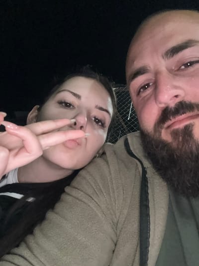 A selfie photograph shows two people looking down at the camera. The person in the upper portion of the frame is male with a beard. The person in the lower portion of the frame is female making a peace sign. The background is indistinct and dark. The location in the city of Goliam Dol, Bulgaria cannot be determined.