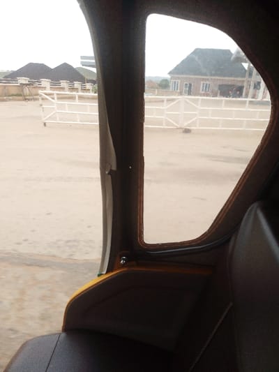The image shows the interior of a yellow and brown tricycle, with the driver's seat to the right of the frame and the side window to the left.  The window is open, showing a view of a dirt road and a white fence in the distance.  There is a building with a gray roof on the other side of the fence. The tricycle appears to be parked near a gas station in Abuja, Federal Capital Territory, Nigeria, at the coordinates provided.  The weather is cloudy, and the road is wet.