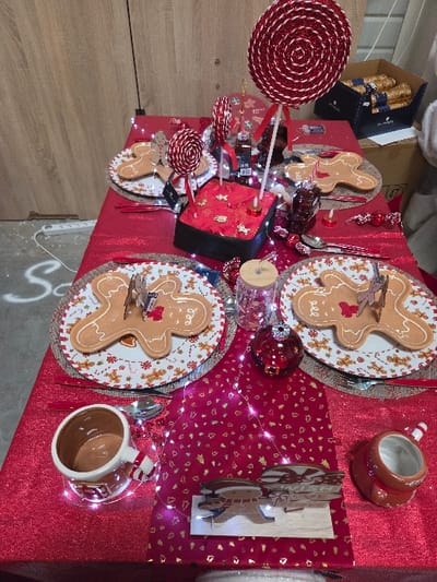 This image depicts a rectangular table meticulously arranged for a festive occasion, located indoors within the city of Blainville-sur-Orne, France. The table is draped with a glittery red tablecloth, overlaid by a narrower red runner patterned with small gold leaf motifs. White string lights are scattered across the table, providing illumination.
Four place settings are visible. Each setting features a round, woven charger plate, topped with a white ceramic dinner plate adorned with a pattern of red and gold gingerbread figures and candy canes. Centered on each dinner plate is a flat, light brown, ceramic gingerbread man-shaped dish, some featuring small red bow details. Red-handled cutlery, including forks, spoons, and knives, is positioned at each setting. Small red candles in metallic holders are interspersed among the settings.
The central part of the table is occupied by a black, rectangular box-like object covered in red fabric, possibly silk, embroidered with gold gingerbread figures. Two small red candles are placed on top of this object. Several decorative red and white spiral lollipops, some glittery, stand upright on white sticks around the central object. A prominent large lollipop decoration is visible on the far right of the table's surface, with smaller ones also present. Additional small red bows, miniature gift box ornaments, and clear glass votive holders or small tumblers serve as accent decorations. A single red Christmas bauble rests on the table's surface towards the lower right. Towards the bottom center, a wooden cutout display features gingerbread figures and the text "WE WISH YOU A MERRY CHRISTMAS" on a stand, illuminated by integrated lights. Two ceramic mugs, designed to resemble gingerbread houses with candy cane-striped handles, are positioned at the bottom left and right of the table.
In the background to the left, a light-toned wooden panel wall or cabinet is visible. A white power strip with a white cable forming an "S" shape is on the floor. To the right, a cardboard box labeled "47" and an open black box containing multiple individually wrapped, gold-colored items are present. A portion of a soft, white object or decoration is partially visible on the far right edge of the frame. No people are present or engaged in any actions.