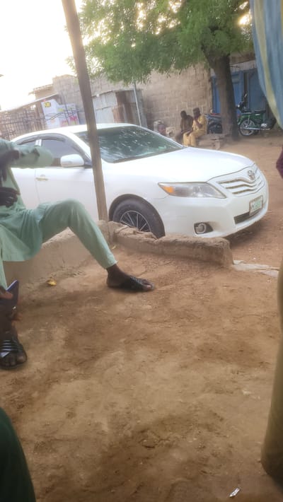 The scene appears to be located in Jogana, Nigeria, featuring a white car parked on a dirt or sandy area adjacent to a building or wall. A person wearing light-colored trousers and black sandals is seen partially within the frame. In the background, there are individuals wearing what appears to be uniforms, possibly school uniforms, standing near a building. A motorcycle is visible behind the car. The setting suggests an outdoor environment within the city.
