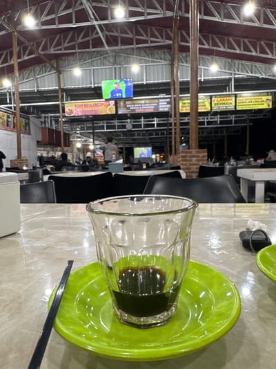 The image depicts a close-up view of a glass filled with a dark liquid, likely coffee, placed on a bright green saucer. A black straw or stirrer rests on the table next to the saucer. The table surface appears to be marble or a similar patterned material. In the background, the scene is set in an open-air food court or restaurant with a metal roof structure supported by columns. Several tables and chairs are visible, occupied by people. Two large television screens are mounted on the ceiling, displaying content, including a sports broadcast on one and what appears to be advertisements or information on others. The overall lighting suggests it is evening or nighttime, with artificial lights illuminating the space. The environment is casual and public, typical of a food establishment in Kalalengkio, Indonesia.