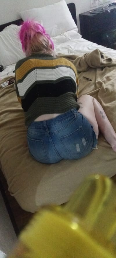 A person with pink hair is sitting on a bed covered in a beige comforter. The person is wearing a striped sweater and denim shorts. The location is Fort Smith, United States of America. A nightstand is visible in the background.