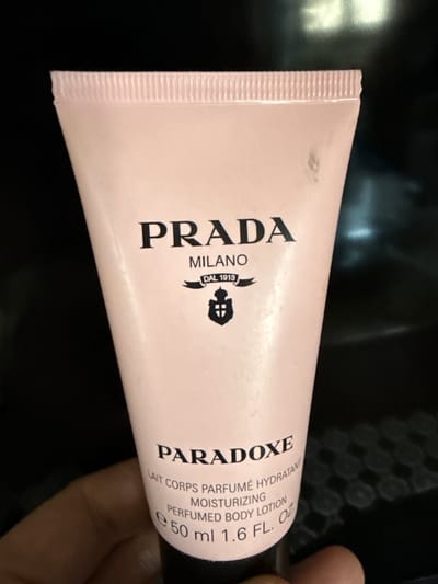 A close-up, angled shot features a pink tube of Prada Paradoxe body lotion, held by a person's fingers against a dark, slightly out-of-focus background. The tube is oriented horizontally, with the cap on the right side. The brand name "PRADA" is prominently displayed in large, black, serif font, followed by "MILANO DAL 1913" and a small emblem. To the right, the product name "PARADOXE" is also in large black font. Smaller text on the tube includes "LAIT CORPS PARFUMÉ HYDRATANT MOISTURIZING PERFUMED BODY LOTION" and "50 ml 1.6 FL. OZ". The lighting suggests indoor conditions, possibly in the evening, with a slightly reflective surface in the background. The focus is on the product, with the person's hand only partially visible, providing a sense of scale and context.