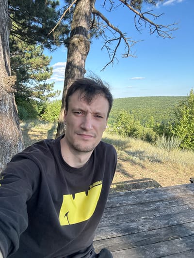 The media file is a selfie taken outdoors. The main subject is a man with short brown hair and a short beard, wearing a black long-sleeved shirt with a large yellow graphic of a smiley face. The shirt also has some yellow text above the graphic, partially visible. He is looking directly at the camera with a neutral expression.
The setting appears to be a natural environment, likely a hilly or mountainous area, based on the background. In the distance, a vast expanse of green forest stretches across the landscape under a bright blue sky with scattered white clouds. The foreground shows dry, yellowish grass and the rough texture of a tree trunk and a wooden surface, possibly a picnic table or bench, which suggests a recreational area within a natural park or forest. The overall lighting indicates it is daytime, likely late morning or afternoon, with clear weather. The image has been rotated, making the man appear to be leaning or lying down. The location is Velykozalissia, Ukraine.