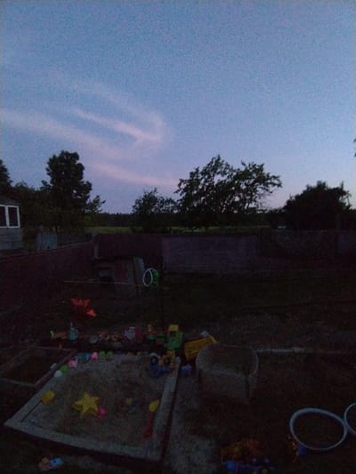 The image captures a twilight scene in a rural backyard, likely in the Talachynski Sel'ski Savet region of Belarus. The sky is a soft, dusky blue, with faint wisps of clouds tinged with pink, suggesting the sun has recently set. The overall mood is peaceful and quiet, characteristic of early evening.
The foreground is dominated by a sandpit filled with various children's toys, including colorful plastic molds and miniature vehicles. A large yellow star-shaped mold is prominent in the sandpit. Scattered around the sandpit are more toys, some appearing to be toy trucks and construction vehicles. The presence of these toys indicates a family setting, likely with young children.
Behind the sandpit, the yard transitions to darker areas, where the ground appears to be grass or dirt. Several indistinct objects are visible, including a dark, possibly wooden structure and a coiled green hose. Further back, a fence or low wall runs horizontally across the frame. Beyond this, the silhouette of trees and a building, possibly a house or shed, can be discerned against the fading light. A dark, wooded area forms the backdrop, suggesting a rural or semi-rural environment.
The lighting conditions are dim, indicating it is dusk. There are no visible people in the frame. The overall impression is one of domestic tranquility, with the day's activities winding down as night approaches.