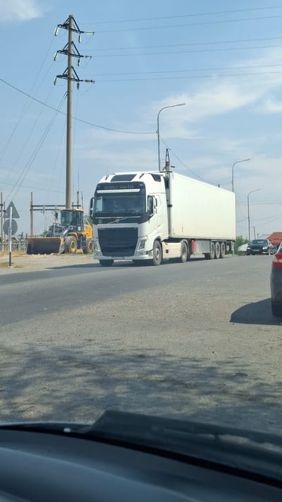 A white Volvo semi-truck with a white trailer is driving on a paved road in an industrial area. The truck has a license plate that reads "748 AQO 13". In the background, there is a yellow bulldozer, power lines, and a few buildings. The sky is blue with some clouds. The image appears to have been taken from inside another vehicle, as the dashboard and windshield are visible in the foreground. The location is identified as Қасымбек датқа, Kazakhstan.