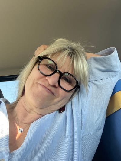 A blonde female is depicted in a close-up, selfie-style photograph taken inside what appears to be a vehicle. She wears dark-rimmed eyeglasses and a light blue, vertically striped collared shirt. A silver-colored chain necklace with a circular, possibly jeweled pendant, is visible on her chest. A dark, rectangular earring is present on her left ear. Her head is tilted, and her right arm is raised, suggesting she is resting her head on her arm or shoulder. Her expression shows a slight, closed-mouth smile. The background is blurred, indicating an indoor setting, likely a car interior, with a bright light source, consistent with daylight, entering from the left. A glimpse of what appears to be a blue and yellow striped object is visible behind her right shoulder. The specific city location of Étoile-sur-Rhône, France, cannot be confirmed from the visual information presented in the image.