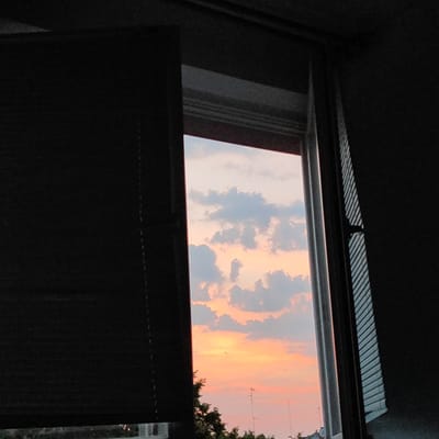 The image shows a view through a window looking out at a sunset. The sky is filled with clouds that are illuminated with shades of orange, pink, and blue. In the foreground, the window frame is visible, with blinds on the left side of the frame. The blinds are dark and partially obscuring the view. The right side of the frame shows a partially closed blind with diagonal lines. The bottom of the image shows a glimpse of trees and some distant structures, possibly buildings and antennas, suggesting an urban or semi-urban setting. The image does not provide any specific details to identify the location as Płock, Poland. There are no people or discernible actions in the scene. The overall impression is one of a calm and serene evening.