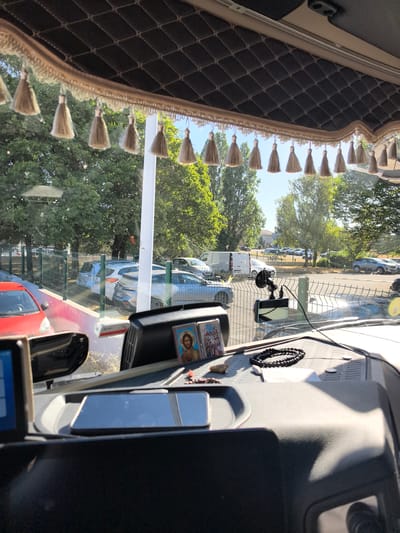 The image is a view from inside a vehicle, likely a truck cab, looking out through the windshield. The interior features a dark, quilted headliner with decorative tassels hanging down. On the dashboard, there is a religious icon with an image of Jesus Christ, a string of black beads, a small stone, and what appears to be a smartphone or GPS device. A dashcam is visible attached to the windshield.
Outside, the scene depicts a parking lot or roadside area with several cars parked behind a metal fence. Trees and greenery form the background, suggesting an outdoor setting in a somewhat urban or suburban environment. The sky is bright and clear, indicating daytime. The presence of parked vehicles and a fence suggests a public or private lot. The overall lighting suggests a sunny day.
The location is identified as Saint-Herblain, France. The specific area within Saint-Herblain appears to be a commercial or industrial zone, given the presence of parked vehicles, including what looks like a white van, and the general landscaping. The fence and the type of vehicles visible do not immediately point to a specific landmark, but the context suggests a functional area rather than a purely residential or tourist spot.