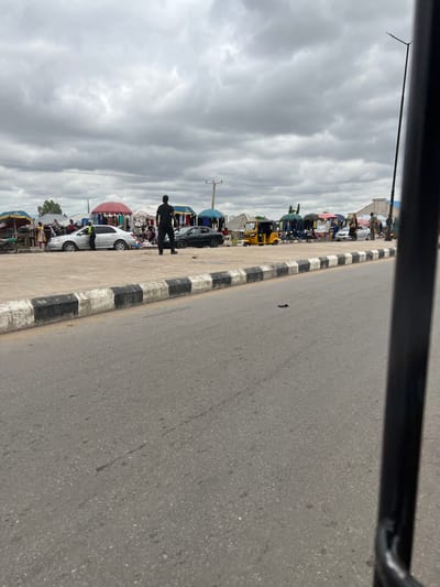 The image depicts a bustling market scene in Gulum, Nigeria. The foreground shows a paved road with a black and white striped curb. In the background, a market is set up with various stalls covered by colorful umbrellas. Several people are visible, some browsing merchandise and others engaged in conversation. Vehicles, including a yellow auto-rickshaw and at least two cars, are present in the market area. A man in dark clothing stands in the middle of the road, observing the market. The sky is overcast, suggesting a cloudy day. The overall impression is one of a lively and active marketplace.