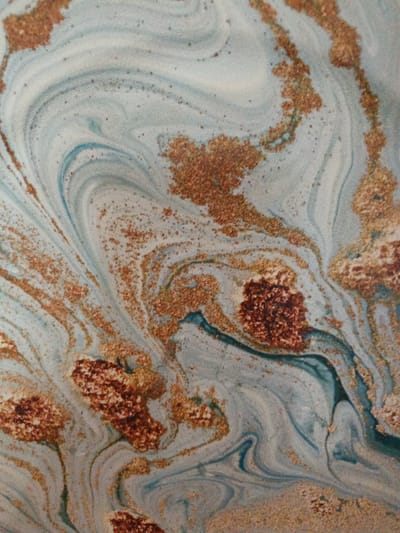 This image shows an abstract, marbled pattern with swirling lines of pale blue, white, and dark teal, interspersed with flecks and larger clusters of a reddish-brown, granular material that appears to be metallic or mineral in nature, possibly gold-colored. The pattern is reminiscent of agate or a similar geological formation, or perhaps a decorative marbling technique on a surface. There are no discernible people, objects, actions, or environmental features of a city or any specific location like Ust-Luga, Russia. The image is a close-up view of a textured surface with an abstract design.
