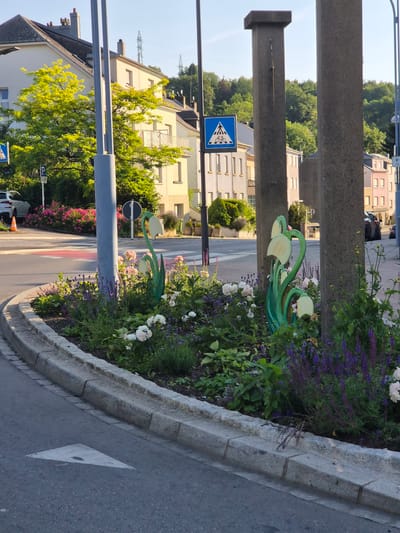The image depicts a street corner in Sanem, Luxembourg, with a flowerbed in the foreground. The flowerbed features green, stylized flower sculptures, purple and white flowers, and green foliage. In the background, there are buildings, trees, and a street with a pedestrian crossing. A traffic sign indicating a pedestrian crossing is visible. The lighting suggests it is daytime, with sunlight casting shadows. The scene appears to be a public space within a town or city.