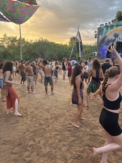 The image displays an outdoor gathering, likely a music event, located in Ban Mae Ping, Thailand, during late afternoon or early evening. Approximately 30 to 40 individuals, primarily young adults, are dispersed across a reddish-brown sandy ground, with many dancing barefoot. In the immediate foreground, a woman wearing a black crop top and shorts is depicted in motion, her right arm raised. To her left, another woman in a green top, a brown satchel, and a rust-colored long skirt dances. Several shirtless men wearing shorts are visible dancing in the mid-ground. Above the scene on the left, a large, multi-colored fabric canopy with spiral and geometric patterns is suspended. On the right, a multi-tiered outdoor stage is visible, equipped with lighting fixtures and a prominent, textured, crescent-shaped decorative element that suggests a moon. A bamboo tripod structure supports an audio speaker positioned near the stage. The background features dense green foliage, with distant hills under a partly cloudy sky illuminated by a golden-orange light from the setting sun.