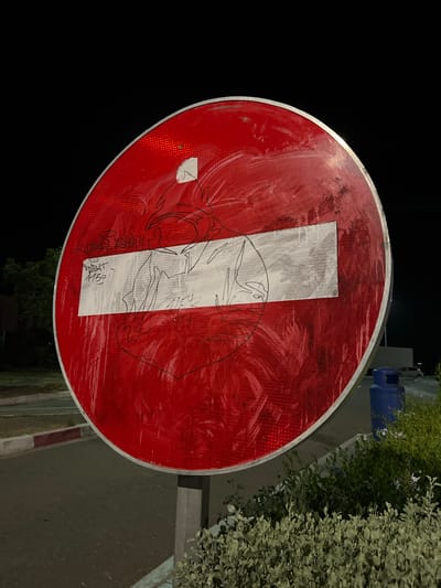 A vandalized "Do Not Enter" sign stands on a street in Jbilate, Morocco. The sign is circular with a red background and a white horizontal bar, partially obscured by white paint and black marker graffiti. The background is dark, suggesting nighttime. A sidewalk and vegetation are visible on the right side of the frame. A blue container sits on the sidewalk.