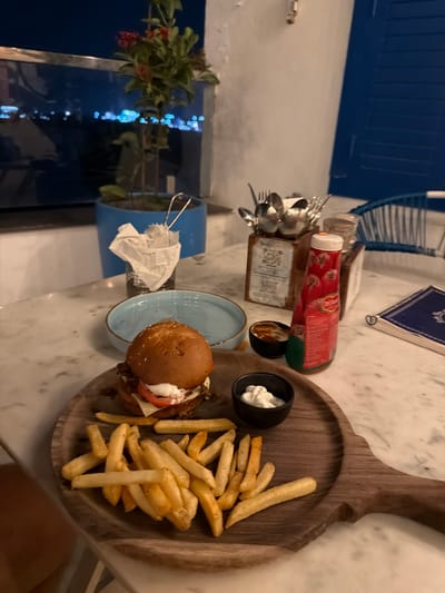 An overhead shot captures a meal setting at night, likely a restaurant, in Shamshabad, India. A wooden board displays a hamburger with lettuce, tomato, and cheese, accompanied by a generous portion of french fries. Two small black bowls hold dipping sauces; one contains a white creamy sauce, and the other, a darker sauce. A bottle of ketchup, possibly Del Monte brand, stands upright next to the bowls. In the background, a blue shuttered window overlooks a nighttime city view with blurred lights, indicating an urban environment. A plant in a blue pot sits near the window, and a wire basket holds crumpled white napkins. Utensils are neatly arranged in a wooden caddy on the marble-topped table. The overall ambiance suggests a casual dining experience with a scenic backdrop.