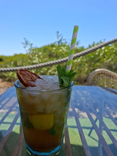 A tall glass filled with ice, a yellow beverage, and mint leaves sits on a glass table. A green and white striped straw is inserted into the drink, and a slice of dried orange is placed on the rim. The drink is garnished with fresh mint leaves and a slice of dehydrated orange. In the background, out-of-focus greenery and a clear blue sky suggest an outdoor setting, likely during the day. The reflection on the table indicates a covered or shaded area, possibly a patio or balcony. The overall impression is one of a refreshing drink enjoyed in a warm, sunny environment. The image captures a moment of leisure and relaxation. There is no visible text in the image.