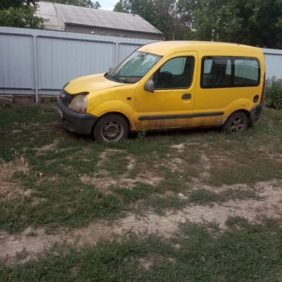 The image shows a yellow van parked on a patch of grass and dirt next to a grey metal fence. In the background, a building with a corrugated roof is visible, along with trees. The van appears to be older and shows signs of wear, with some dirt and rust on its exterior. The area surrounding the van is overgrown with grass. The location is identified as Slobidka, Ukraine.