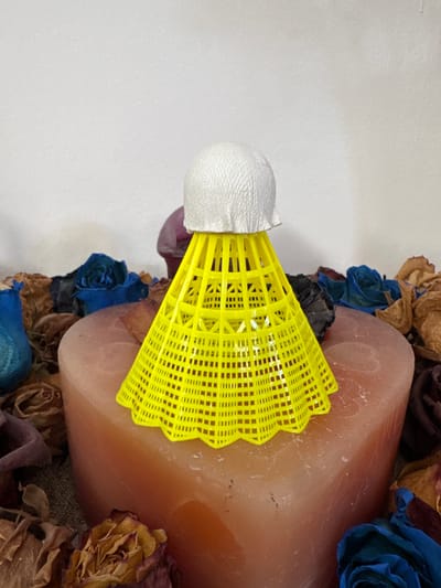 The image displays a close-up of an artistic arrangement that includes a badminton shuttlecock, a large cylindrical candle, and dried flowers. The shuttlecock, with its white feathered end and bright yellow plastic skirt, is positioned horizontally, leaning against the candle. The candle itself is substantial in size and has a smooth, opaque surface with a peachy-pink hue. Surrounding the candle and shuttlecock is an assortment of dried flowers and petals. These include roses in various shades of blue, brown, and purple, some appearing desiccated and delicate, others more vibrantly colored. The background is a plain, textured white surface, suggesting it might be a wall or backdrop. The lighting is soft and even, creating a studio-like atmosphere. There is no visible text within the image, and no indication of a specific time of day, weather, or overt emotional cues, other than the somber tone often associated with dried flowers. The composition focuses on the textures and colors of these disparate elements, creating a still-life arrangement. The location context of Sant'Angelo di Piove di Sacco, Italy, is provided externally and not discernible from the image itself.