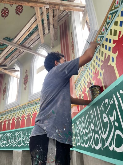 Inside a building in Mns Asan, Indonesia, a male painter is depicted from the back and right side, actively decorating a wall. He wears a paint-splattered dark grey t-shirt and dark pants. His right hand holds a thin paintbrush, meticulously applying light blue paint to an ornate yellow and red patterned section of the wall, positioned below an arched window. His left hand holds a small, dark, paint-splattered bucket.
The wall features multi-colored Islamic art: a prominent lower green band displays white Arabic calligraphy, surmounted by a red section with yellow geometric and floral patterns. Further up, pale olive-green walls exhibit additional red decorative motifs. Multiple arched windows, letting in bright natural light, are visible. Overhead, crude wooden scaffolding is present, indicating ongoing construction or renovation of the interior space.