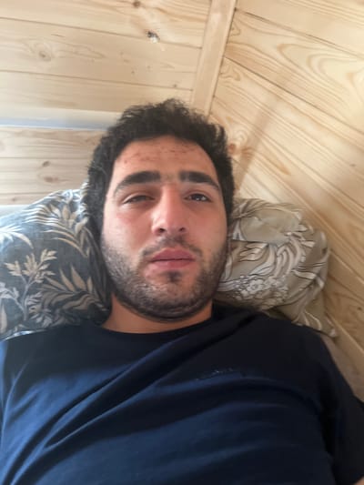 The image is a first-person perspective selfie of a young man lying down on a bed. He has dark, curly hair and a beard, and appears to have some acne on his forehead and cheeks. He is wearing a dark blue t-shirt. He is lying on two pillows, one with a blue and white floral pattern and the other with a light brown and white pattern. The background consists of wooden walls, likely part of a room with an angled ceiling. The lighting is natural and seems to be coming from above. There is no indication of the specific location within Sergeevka, Russia.