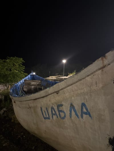 The image shows a close-up, low-angle view of the side of a white and blue boat at night, resting on land, with the name "ШАБЛА" (Shabla) painted in blue on its hull. The boat appears weathered and worn, with visible paint peeling and rust stains. The interior of the boat, visible at the front, shows blue paint and wooden seating. In the background, a modern streetlamp illuminates the scene, casting light on trees and vegetation. The sky is dark, indicating nighttime. The boat is located in the city of Tyulenovo, Bulgaria.