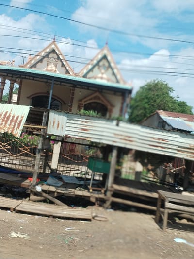 Thôn 4, Vietnam. A two-story building with a gabled roof is visible in the background. In the foreground, a corrugated metal structure covers an area with wooden planks and debris scattered on the ground. A rusty metal fence is present. Overhead, power lines cross the sky with blue sky and white clouds.