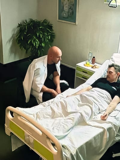 The image depicts an indoor scene, likely a hospital room, where two adult males are present. One male, who appears to be a medical professional due to his white lab coat, is kneeling beside a hospital bed. He has a shaved head and is looking down at the person in the bed. The other male is lying in the hospital bed, covered by white sheets. He is wearing a black t-shirt and has short, dark hair. An IV line is visible on his arm.
In the background, there is a bedside table with various items, including a bottle of water and some food. A potted plant with green leaves is situated to the left of the scene, and a framed picture hangs on the wall above the bedside table. The overall environment suggests a medical setting. The location is identified as Dondușeni, Moldova.