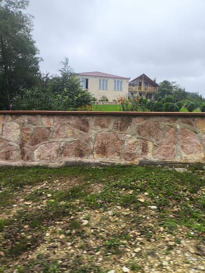 The image depicts a natural scene with two houses in the background. The foreground is dominated by a stone wall constructed with irregular, large stones of reddish-brown hues, mortared together with grey cement. A dark reddish-brown coping caps the wall. In front of the wall, there is a patch of green grass interspersed with small pebbles and some fallen leaves, suggesting a somewhat unkempt area. Behind the wall, lush green foliage, including bushes and trees, is visible.

Further in the background, two houses are situated on a slight incline. The house on the left is a light-colored, two-story structure with a reddish-brown roof and white-framed windows. In front of this house, there is a garden area with blooming flowers in yellow and orange, along with some purple flowers further to the left. The house on the right is a multi-story wooden house with a steeply pitched roof and a large balcony. Stairs lead up to the balcony. The houses are surrounded by more greenery and trees. The sky above is overcast with grey clouds. The overall impression is that of a residential area nestled within a natural, somewhat rural or suburban environment, likely in Skurdi, Georgia.