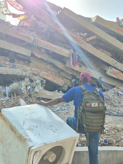 In Caraballeda, Venezuela, a man wearing a blue shirt, jeans, and a red cap stands in front of a collapsed building, with his hand resting on a damaged washing machine. He is equipped with a green backpack adorned with patches, including one depicting the Venezuelan flag and another with "161 ANIVERSARIO". Behind him, the immense debris of the destroyed structure looms, with some flags and clothing items visible hanging precariously from the wreckage. Other individuals can be seen in the background amidst the rubble, suggesting ongoing search or recovery efforts. The environment is characterized by destruction and debris, indicative of a catastrophic event.