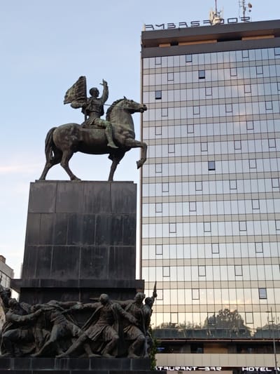 The image depicts a bronze equestrian statue of a figure holding a flag and pointing with an outstretched arm, positioned in front of a modern glass-facade building. Below the equestrian statue, there is a relief sculpture of several figures. The building features numerous square windows and a sign at the top that reads "AMBASADOR." The lower part of the building's facade has a sign that appears to read "RESTAURANT - BAR" and "HOTEL". The scene is lit by natural light, suggesting it is daytime, with the sky visible and reflecting in the building's windows. This statue is the "Partisans Liberation Monument" in Niš, Serbia, located in front of the Hotel Ambasador.