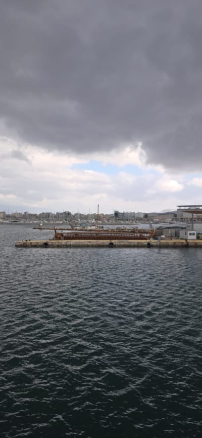 The image presents a panoramic view of a harbor or port city under a dramatic, partially cloudy sky. The perspective is from an elevated position, looking across the water towards the distant city.

The upper portion of the image is dominated by the sky. On the left, dark, heavy grey clouds are prominent, suggesting overcast weather or an impending storm. Towards the center and right, the cloud cover lightens significantly, revealing patches of pale blue sky peeking through, indicating a break in the clouds.

The lower right and central-right sections of the image are filled with water. The water appears dark blue-green and is covered with numerous small ripples and waves, suggesting the presence of a breeze or current. The texture of the water surface is well-defined by the interplay of light and shadow from the overcast sky.

Stretching horizontally across the midground is a long, narrow pier or dock. It appears to be made of concrete or stone, with a somewhat weathered, light brown or reddish-brown hue visible on some structures along its length, possibly indicating rust or aged materials. Several dark, cylindrical bollards are evenly spaced along the pier's edge, likely used for mooring vessels.

Beyond the pier and across the water, a sprawling city or town lines the distant shoreline. Numerous buildings of various sizes and architectural styles create a dense urban landscape. Many tall, slender masts of boats are visible clustered along the waterfront, suggesting an active marina or anchorage. A tall, crane-like structure can be faintly seen among the buildings, indicating industrial activity within the port. In the far distance, subtle outlines of mountains or hills rise behind the city on the left.

The overall lighting is diffuse and soft, consistent with an overcast day. The scene captures a dynamic weather condition, with both dark, heavy clouds and brighter patches of sky, creating a moody yet expansive atmosphere. There is no visible text or identifiable people in the image.