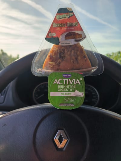 This media file shows a close-up view of a car's steering wheel with two food items placed on top of it. The steering wheel has the Renault logo in the center. Above the steering wheel, a triangular plastic container holds a slice of banana bread topped with nuts. The packaging for the banana bread has a label that reads "Becky's Factory BANANA BREAD Avec éclats de noix" and "Fabriqué en France". Below the banana bread is a small, round green container of Activia yogurt. The label on the yogurt reads "DANONE ACTIVIA BIEN-ÊTRE DIGESTIF NATURE" with a best-before date of "01/07 25". The scene appears to be outdoors, with a bright sky and some greenery visible in the background, suggesting it is daytime and the weather is fair. The overall impression is a quick snack or meal being consumed inside a parked car.