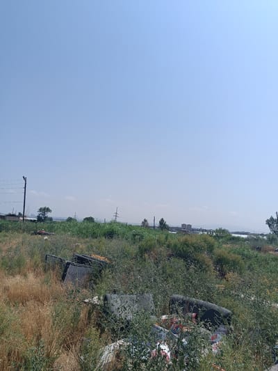 This is a wide-angle, eye-level, outdoor photograph taken on a clear, sunny day in Ghukasavan, Armenia. The foreground is filled with overgrown dry grasses and dense green bushes. Amongst the foliage, several discarded objects are visible. In the mid-ground, there are what appear to be large, black, irregular-shaped pieces of debris, possibly furniture or industrial waste. Some colorful plastic containers are also scattered within the vegetation.

Further back, a vast expanse of green vegetation stretches across the landscape. In the distance, a hazy skyline is visible, featuring a body of water, distant buildings, and structures that suggest an urban or industrial area. Several power poles and electrical lines are also present, traversing the horizon. The sky is a pale, cloudless blue. The scene depicts an area with significant overgrowth and the presence of accumulated waste, set against a backdrop of a distant town or city.