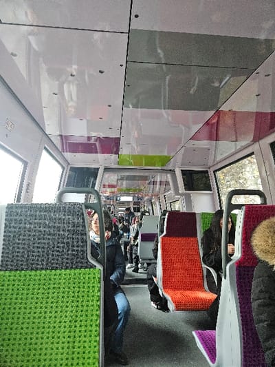 The image depicts the interior of a public transport passenger carriage, located in the city of Levallois-Perret, France. The perspective is from a slightly elevated position, looking forward down a central aisle.
Multiple passengers are present. In the left mid-ground, a person wearing a dark blue jacket, blue jeans, and a light-colored scarf is seated, partially obscured by a seat in front, looking towards the right side of the carriage. In the right mid-ground, the back of a person's head and a dark jacket with a light, possibly faux-fur, collar are visible. Further down the right side, a person with long dark hair is seated, looking downwards. Other indistinct figures are visible further down the aisle, both seated and possibly standing.
The carriage features modern, individual bucket-style seats upholstered in a textured fabric. Seat colors vary, displaying sections of dark grey, bright green, orange, and purple/burgundy. The seat in the immediate foreground on the left is grey on top and bright green below. A seat on the right foreground is purple/burgundy on top and purple below. Grey metal handrails are integrated into the top of the seatbacks. Large rectangular windows line both sides, revealing a blurred exterior consistent with motion, showing trees and an overcast sky. The ceiling consists of light-colored, reflective panels interspersed with darker grey rectangular sections, secured by visible fasteners. The floor is dark grey and textured. A small white square emergency pictogram sign is visible on the upper left wall. The interior appears clean and well-maintained.