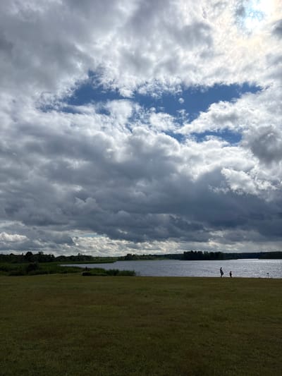 The image shows a wide, grassy field in front of a large body of water. A treeline borders the far side of the water. The sky is dramatic, with large, dark clouds mixed with patches of blue sky and bright sunlight. Two silhouetted figures, one adult and one child, are walking near the water's edge in the middle ground. The scene is likely an outdoor recreational area in Мазалаўскі сельскі Савет, Belarus.