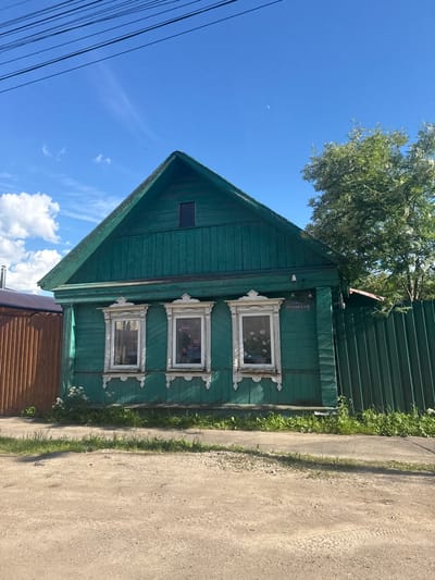 A bright green, wooden, one-story house with a dark green pitched roof sits on a dirt road in Vereya, Russia. The house has three white-trimmed windows with ornate white wooden carvings, and the windows appear to contain potted plants. A sign above the rightmost window reads "УЛИЦА ЛЕНИНСКАЯ" (Leninskaya Street). A green metal fence runs along the right side of the house, and a wooden fence is visible to the left. A large green tree is behind the house and to the right. The sky is blue with scattered white clouds and a sliver of the moon is visible. Power lines are strung across the top of the frame.