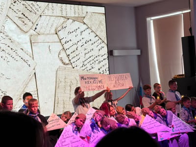 A group of children, some with face paint and blue and white scarves, are on a stage in what appears to be a school auditorium in Pron'kino, Russia. They are holding signs and some are holding candles. A large projection screen behind them displays old letters. Two prominent signs held by older children read "WE REMEMBER EVERYONE WHO DID NOT RETURN" and "RETURN HOME". The children are participating in an event that appears to be a memorial or a patriotic ceremony. The lighting suggests an indoor performance space.