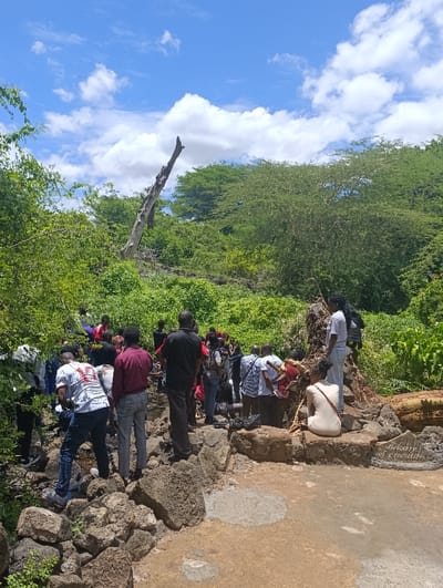 A group of approximately twenty people are gathered in a rocky area near a dry riverbed. The location is near the coordinates -2.984 latitude and 38.023 longitude, which is located near the Tsavo East National Park in Kenya.  The group is comprised of mostly men, with a few women scattered throughout.  A woman in a white dress sits on a rock with her legs crossed.  A few people are standing in a small group, facing the left, while the majority of the people are facing right and appear to be in conversation or looking at something to the right of the image. The people are all wearing casual clothing. There are a few trees and shrubs visible in the background, as well as a large, dead tree that is leaning to the right.  The sky is blue and contains several white, fluffy clouds. The ground is dry and rocky.  A sign made from a flat rock reads "Welcome to the Cradle".