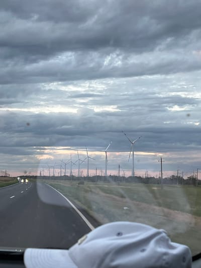 The image shows a landscape scene dominated by a cloudy sky and a field with wind turbines. The perspective suggests the photo was taken from a vehicle, with a portion of a white cap visible in the foreground, obscuring the right side of the frame.
The main subjects are the numerous wind turbines spread across the field, silhouetted against the sky. Some of the turbines are in motion, with their blades extended. Power lines and poles are also visible, running parallel to the wind turbines. The ground appears to be a grassy field, with a paved road or pathway curving through the scene. In the distance, a line of trees can be seen.
The time of day appears to be either dawn or dusk, given the soft lighting and the colors in the sky, which range from dark grays to lighter blues and pinkish hues. The weather is overcast, with thick clouds covering most of the sky.
The location cues, such as the wind turbines and the rural landscape, are consistent with a power generation facility in an open area. There is no visible text in the image. The overall mood conveyed is one of a vast, industrial landscape under a dramatic sky. There is no clear indication of specific activity or event beyond the implied function of the wind turbines.