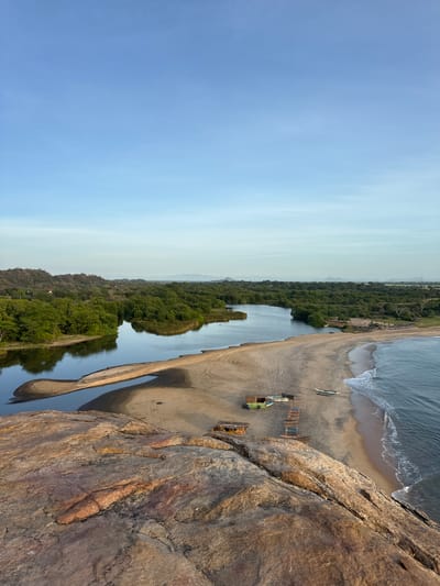 This is a landscape photograph taken from a high vantage point, likely a cliff overlooking a coastal area. The scene depicts a bay where a calm, dark body of water, possibly a river or inlet, meets the sandy shore of the ocean. Gentle waves roll onto the beach from the deep blue ocean. 

On the sandy beach, there are several objects suggesting human activity. Two long, narrow boats, likely canoes or kayaks, lie on the sand. Further inland, a small, rustic wooden structure with a thatched roof is visible, possibly a shelter or stall. A green vehicle, perhaps a small truck or jeep, is parked near this structure, alongside more wooden seating arrangements. The beach is bordered by a dense, lush green forest that extends into the distance. Beyond the forest, rolling hills and mountains are faintly visible in the hazy distance.

The photograph is captured in portrait orientation, with the sky occupying the left third of the image. The sky is a soft, pale blue, transitioning to a lighter shade at the horizon, suggesting either early morning or late afternoon. The overall mood is tranquil and serene, highlighting the natural beauty of the coastal landscape. No people are visible in the frame, and there is no visible text. The location context is Ulla, Sri Lanka.