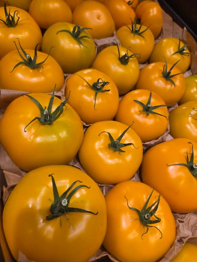 A top-down, close-up shot shows a bulk display of ripe, yellow tomatoes. The tomatoes are mostly round with a smooth, glossy skin, and have green stems and leafy calyxes. They are arranged in several layers in what appears to be a cardboard container, suggesting a market or grocery store setting. The lighting is bright, highlighting the vibrant color of the tomatoes and creating subtle reflections on their surfaces. The background consists of more tomatoes and brown paper material. There are no people or other objects clearly visible in the frame. The scene is entirely focused on the produce.