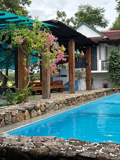 This is a medium shot, eye-level, outdoor, daytime, full-color photograph taken in Bố Trạch Commune, Vietnam. The image depicts a poolside scene at a resort or restaurant. In the foreground, a bright blue swimming pool occupies the bottom right half of the frame, bordered by a stone wall. The wall is constructed with larger, rough-hewn stones on the bottom, topped with a band of smaller, smooth white pebbles. A chain-link fence is visible along the pool's edge, and ripples are present on the water's surface.
To the left of the pool, the stone wall continues, and behind it is a covered outdoor seating area. This area features a roof made of turquoise corrugated plastic supported by bamboo poles. A vibrant pink bougainvillea plant with green leaves cascades over a corner of the structure. Wooden benches are visible beneath the overhang, and a bicycle is parked to the far left.
Behind the seating area, a white building with a red tile roof is visible. It has multiple stories and balconies. Several colorful, lantern-like decorative objects hang from the roof of the seating area. A standing fan is positioned near the building. The background consists of trees and a cloudy sky. There are no people visible in the image.