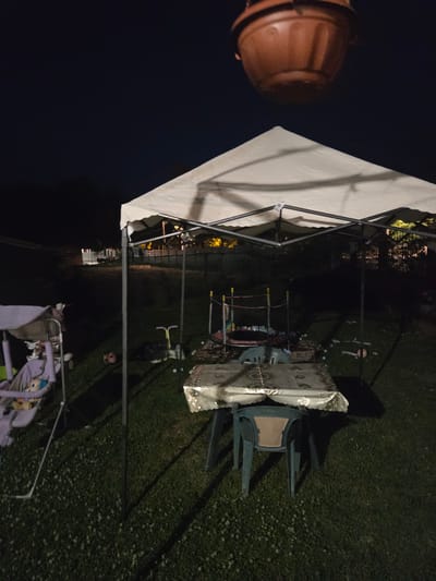 The image depicts an outdoor scene at night, possibly in a backyard. A white canopy tent is set up, covering a table with a decorative tablecloth and several chairs. To the left, a child's swing set with a purple seat is visible. In the background, there are dimly lit structures and trees. The ground is covered with grass. A brown, hanging planter is suspended in the upper part of the frame. The scene appears to be in Todor Ikonomovo, Bulgaria, based on the prompt's context.