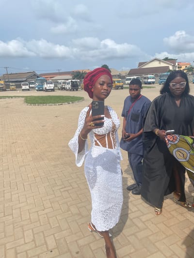 The image depicts four individuals standing on a paved surface. A woman in a white dress with a red head wrap is holding a phone and appears to be taking a selfie. Behind her, there is a person in a blue shirt. The woman in black is holding a phone and a patterned bag. The location appears to be a parking lot or courtyard with buildings and vehicles in the background. The location is Otta, Nigeria.