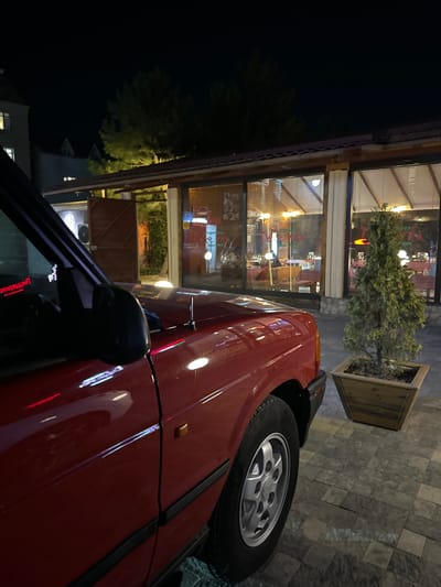 In Orto-Sai, Kyrgyzstan, at night, a red SUV is parked in front of a building with large glass windows, likely a restaurant or cafe. The building's interior is illuminated, revealing tables and chairs. A small evergreen tree in a wooden planter sits to the right of the SUV. The ground is paved with interlocking stone tiles. Reflective lights from the building and possibly streetlights are visible on the car's glossy red paint. The sky is dark, indicating the late hour.