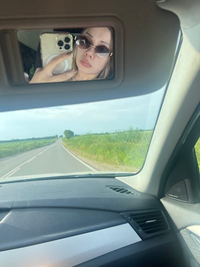 The image displays the interior of a vehicle, likely captured from a forward-facing perspective, overlooking a rural road situated near Stefanovo, Bulgaria.
A female individual is visible in the reflection of the passenger-side vanity mirror, engaged in taking a selfie. She possesses light-colored hair and wears large, rectangular sunglasses with gold-toned accents. Her lips are full, and her fingernails are painted a dark shade of brown or mauve. She holds a gold-colored iPhone, identifiable by its triple-lens camera array and clear case featuring a white MagSafe ring.
The vehicle's dashboard is dark grey with a textured surface, accentuated by a horizontal silver trim panel on the passenger side. A passenger-side air vent, equipped with a silver adjustment tab, is visible, alongside an "AIRBAG" label. A Harman Kardon branded speaker grille is located in the upper right portion of the passenger compartment.
Beyond the windshield, a two-lane asphalt road with double solid white center lines extends into the horizon under a light blue sky with minimal cloud cover. Green fields flank both sides of the road, with red flowers, consistent with poppies, sparsely visible within the vegetation on the right side. A cluster of trees is discernible in the distant right background. The scene is consistent with a daytime setting in the region of Stefanovo, Bulgaria.