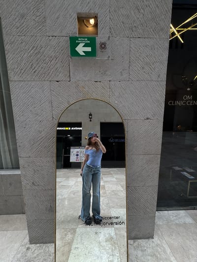 Here is a forensic description of the image:
**Overview:**
The image shows a street scene, possibly in Ciudad López Mateos, Mexico, indicated by text that appears in the reflection. The photo is likely taken by someone looking into a mirror.
**Environment:**
The surrounding environment appears to be a modern urban space with stone cladding on the walls. A sign reading "RUTA DE EVACUACIÓN" (Evacuation Route) with a downward-pointing arrow is mounted on the stone wall, next to a recessed light fixture. A sign for "OM CLINIC CENTER" is visible.
**Subjects and Objects:**
* A person is visible in the mirror, taking a picture of themselves. The person is wearing a light-colored shirt, jeans, and a cap.
* In the background reflected in the mirror, signage for "ИАНОЖЭ AVONNI" and "OM CLINIC" can be seen. A sign with foreign exchange rates is also visible.
**Actions and Interactions:**
The individual in the mirror is using a device, probably a smartphone, to capture a photograph or selfie. The mirror reflects the surrounding urban environment and signage.