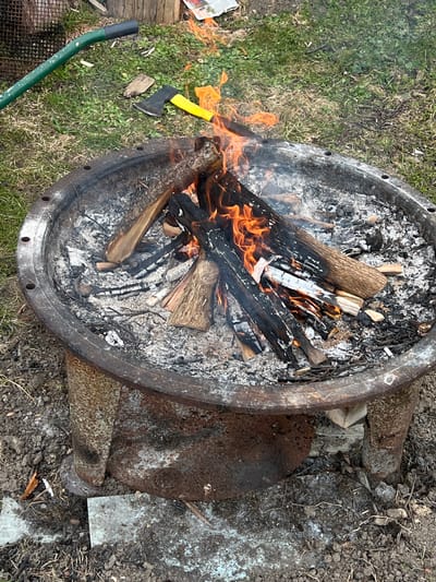 The image depicts a metal fire pit containing burning wood and ash. The pit is located outdoors on a patch of grassy ground interspersed with dirt and debris. To the left of the pit, an axe with a yellow handle rests on the grass. A green metal rod is also visible. The fire pit itself appears to be constructed from heavy metal, showing signs of rust and weathering. It is supported by a rusted metal base. The surrounding environment consists of soil, scattered leaves, and patches of sparse vegetation. The location, Dārzciems, Latvia, is a relevant but not readily apparent detail within the image itself.