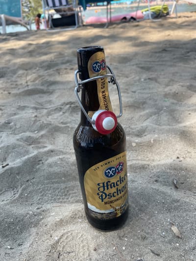 A brown glass bottle of Hacker-Pschorr Münchner Gold beer lies on its side in the sand, with a metal swing-top closure and a red and white stopper. The label is golden with text. Some of the visible text on the label reads "Hacker-Pschorr", "MÜNCHNER GOLD", and "DER HIMMEL DER BAYERN". In the background, out of focus, is a sandy beach with some beach chairs, umbrellas, and people in the distance. The scene suggests a sunny day at the beach.
